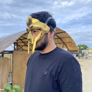 May include: A gold-colored mask with a distinctive design, featuring a prominent faceplate and a headpiece. The mask has a metallic sheen and is worn with a black strap. The person is wearing a dark blue shirt.