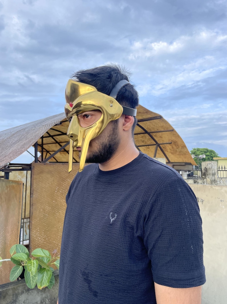 May include: A gold-colored mask with a distinctive design, featuring a prominent faceplate and a headpiece. The mask has a metallic sheen and is worn with a black strap. The person is wearing a dark blue shirt.