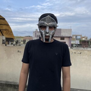 May include: A silver-colored mask with intricate details, featuring a stylized face design. The mask is worn over the face. The person is wearing a black t-shirt.