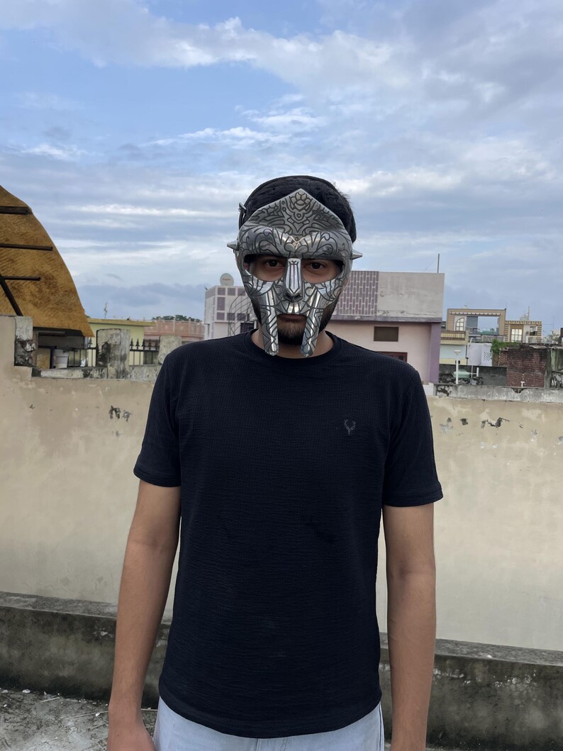 May include: A silver-colored mask with intricate details, featuring a stylized face design. The mask is worn over the face. The person is wearing a black t-shirt.