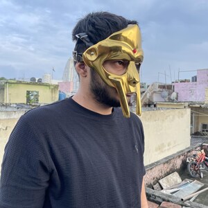 May include: A gold-colored mask with a distinctive design, featuring a prominent nose and a red accent. The mask has a leather strap for securing it. The mask is a unique accessory.