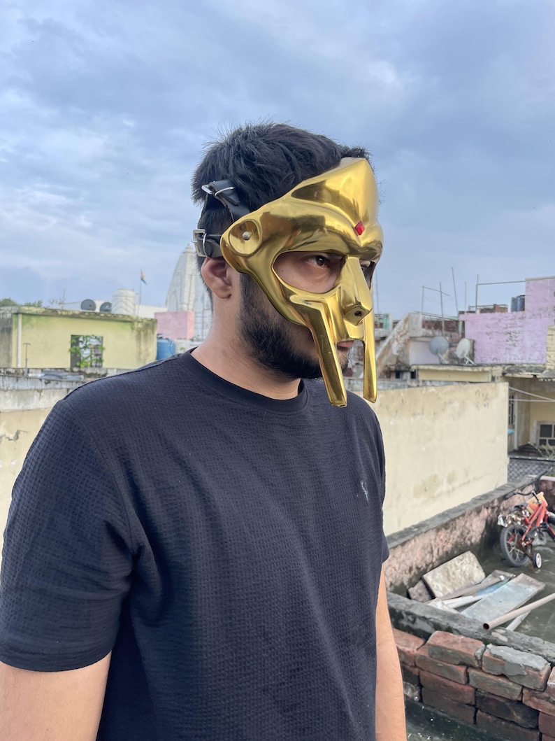 May include: A gold-colored mask with a distinctive design, featuring a prominent nose and a red accent. The mask has a leather strap for securing it. The mask is a unique accessory.