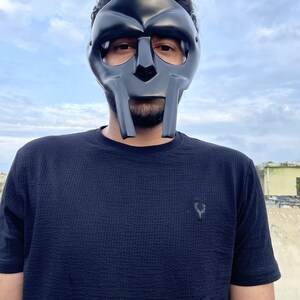 May include: A black mask with a distinctive, angular design, covering the face. The mask has a smooth, matte finish and is worn against a backdrop of a cloudy sky. The person is wearing a black textured t-shirt.