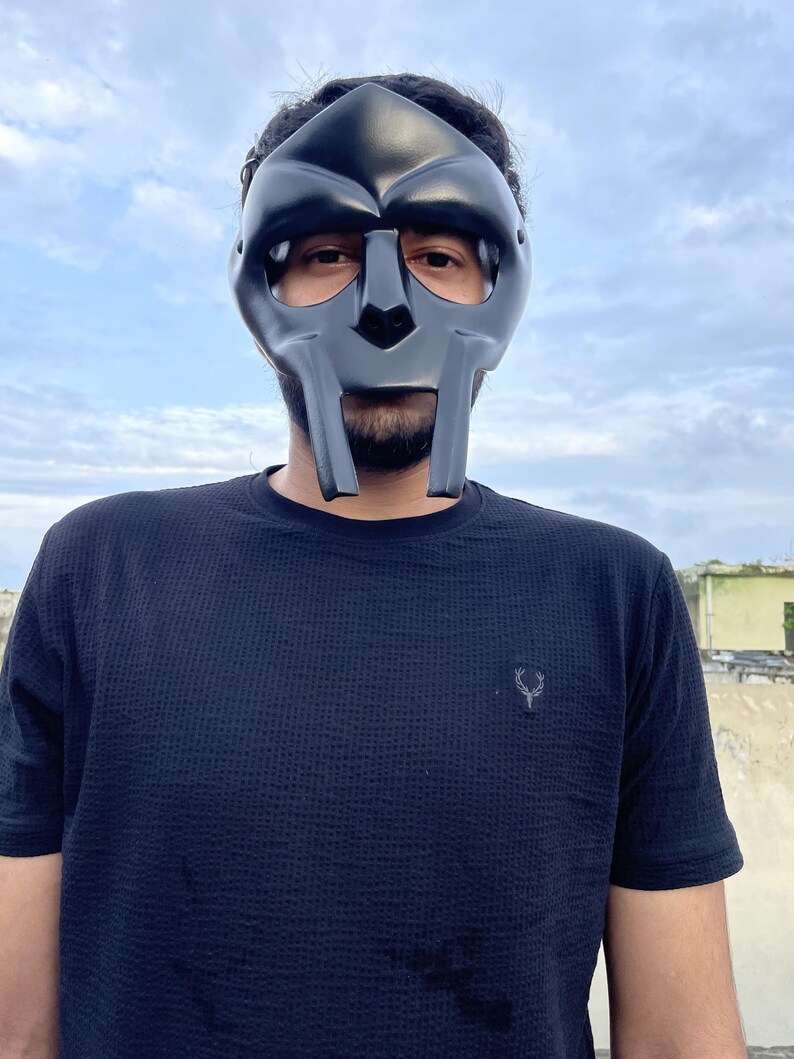 May include: A black mask with a distinctive, angular design, covering the face. The mask has a smooth, matte finish and is worn against a backdrop of a cloudy sky. The person is wearing a black textured t-shirt.
