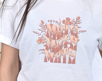 Puede incluir: Camiseta blanca con un diseño floral y el texto "Make your own magic" en color rosa melocotón.