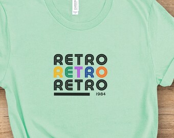 Könnte beinhalten: Ein hellgrünes T-Shirt mit einer schwarzen und bunten Grafik, auf der viermal "Retro" und darunter "1984" steht.