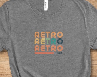 Könnte beinhalten: Ein graues T-Shirt mit einem Retro-Design, das das Wort "Retro" viermal in verschiedenen Farben wiederholt und ein kleines "1984" darunter gedruckt ist.