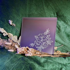 Może przedstawiać: Kwadratowe lustro z fioletowo-różowym gradientem i białym kwiatowym wzorem w rogu. Lustro jest oparte na podłożu z suszonych kwiatów i zieleni na ciemnozielonym tle.