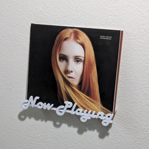 May include: A close-up of a record album cover featuring a woman with long red hair. The album cover is black with white text that reads "Vera Blue Perennial". The text "Now Playing" is written in white on a white background.