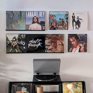 Może przedstawiać: Biała ściana z gramofonem i półką z płytami winylowymi. Płyty należą do takich artystów jak Lana Del Rey, Village People, Fleetwood Mac, The Verve, Daft Punk i Jeff Buckley.