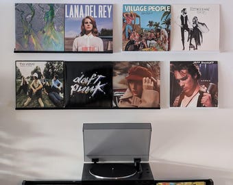 May include: A white wall with a record player and a shelf of vinyl records. The records include artists like Lana Del Rey, Village People, Fleetwood Mac, The Verve, Daft Punk, and Jeff Buckley.