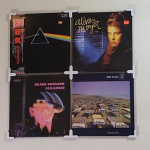 May include: Four vinyl records displayed on a wall with white corner frames. The records are Pink Floyd's "Dark Side of the Moon", Alison Moyet's "Alf", Black Sabbath's "Paranoid", and Pink Floyd's "The Wall".