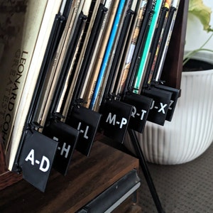 Puede incluir: Un soporte de discos de metal negro con divisores negros etiquetados A-D, E-H, I-L, M-P, Q-T, J-X y Z. El soporte está lleno de discos de vinilo.