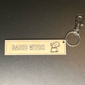 May include: A wooden keychain with a brown background and a white outline. The keychain has the text "BASIC WITCH" and a cartoon image of a cauldron with steam coming out of it.