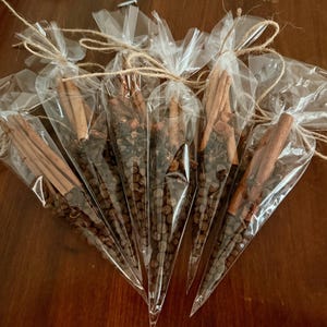 May include: Five clear cellophane bags filled with spices, tied with twine. Each bag contains a mix of whole spices, including cinnamon sticks, peppercorns, and star anise. The bags are arranged on a dark wooden surface.
