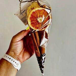 May include: A clear cellophane cone filled with dried orange slices, cinnamon sticks, and coffee beans, tied with twine. A small tag with handwritten text is attached. The arrangement is held in a hand with a white watch.
