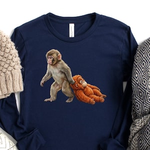 May include: Navy blue long-sleeve shirt featuring a graphic of two monkeys. A tan knit beanie, a decorative pumpkin, and a patterned scarf with cotton stems are also in the image. The items are arranged on a white surface.