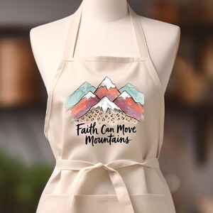 May include: A cream-colored apron with a mountain graphic in shades of red, teal, and white. The apron has a front pocket and a tie-waist. The text "Faith Can Move Mountains" is printed below the mountain graphic.