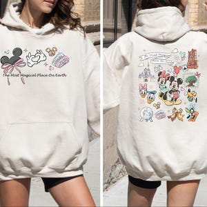 以下が含まれることがあります： 前面にミッキーマウスの耳、ハートを作る手、プレッツェルが描かれたクリーム色のパーカー。「The Most Magical Place On Earth」の文字がプリントされています。パーカーの背面には、さまざまなディズニーのキャラクターやランドマークが描かれています。