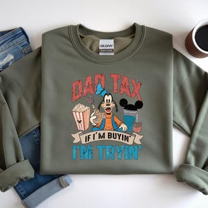 Pode incluir: Uma camisola de gola redonda verde-azeitona com um gr&aacute;fico da personagem Pateta a segurar um pretzel, pipocas e uma bebida. O texto DAD TAX IF I'M BUYIN' I'M TRYIN' est&aacute; em letras vermelhas e azuis desgastadas. Uma etiqueta Gildan &eacute; vis&iacute;vel.