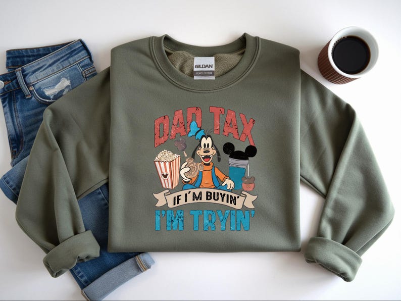 Pode incluir: Uma camisola de gola redonda verde-azeitona com um gr&aacute;fico da personagem Pateta a segurar um pretzel, pipocas e uma bebida. O texto DAD TAX IF I'M BUYIN' I'M TRYIN' est&aacute; em letras vermelhas e azuis desgastadas. Uma etiqueta Gildan &eacute; vis&iacute;vel.
