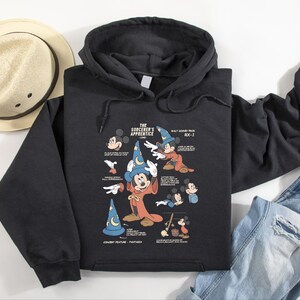 Puede incluir: Una sudadera negra con capucha que presenta un estampado gr&aacute;fico de El Aprendiz de Brujo con Mickey Mouse con un sombrero de mago azul y una t&uacute;nica roja, rodeado de varios bocetos, notas t&eacute;cnicas y el texto Walt Disney Prod RX-1.