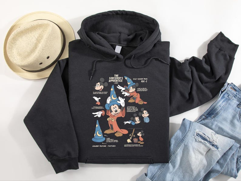 Puede incluir: Una sudadera negra con capucha que presenta un estampado gr&aacute;fico de El Aprendiz de Brujo con Mickey Mouse con un sombrero de mago azul y una t&uacute;nica roja, rodeado de varios bocetos, notas t&eacute;cnicas y el texto Walt Disney Prod RX-1.