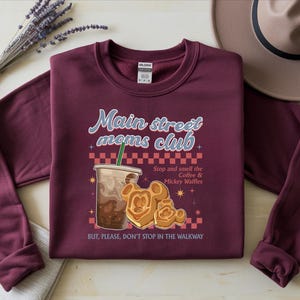 Op de afbeelding: Bordeaux sweatshirt met de tekst "Main street moms club" en een afbeelding van koffie en Mickey Mouse wafels. De tekst "Stop and smell the Coffee & Mickey Waffles" is ook opgenomen. Een hoed en een lavendeltakje staan op de achtergrond.