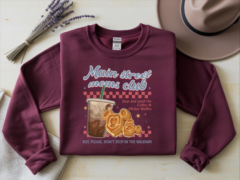 Op de afbeelding: Bordeaux sweatshirt met de tekst "Main street moms club" en een afbeelding van koffie en Mickey Mouse wafels. De tekst "Stop and smell the Coffee & Mickey Waffles" is ook opgenomen. Een hoed en een lavendeltakje staan op de achtergrond.