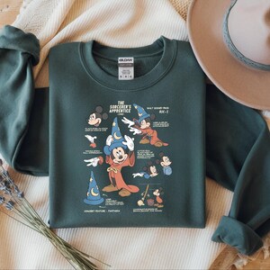 Puede incluir: Una sudadera de cuello redondo en color verde bosque oscuro con un estampado gr&aacute;fico de El Aprendiz de Brujo de Fantas&iacute;a. El dise&ntilde;o incluye varias ilustraciones de Mickey Mouse con sombrero y t&uacute;nica de mago, junto a etiquetas de texto.