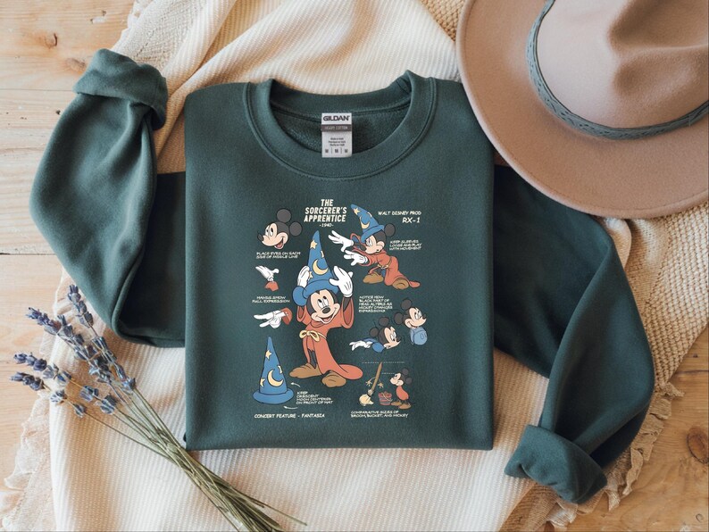 Puede incluir: Una sudadera de cuello redondo en color verde bosque oscuro con un estampado gr&aacute;fico de El Aprendiz de Brujo de Fantas&iacute;a. El dise&ntilde;o incluye varias ilustraciones de Mickey Mouse con sombrero y t&uacute;nica de mago, junto a etiquetas de texto.