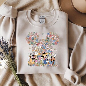 May include: Beige sweatshirt featuring a floral Mickey Mouse design with Mickey, Minnie, Donald, Daisy, Goofy, and Pluto. The design is surrounded by colorful flowers. The sweatshirt has long sleeves and a crew neck.