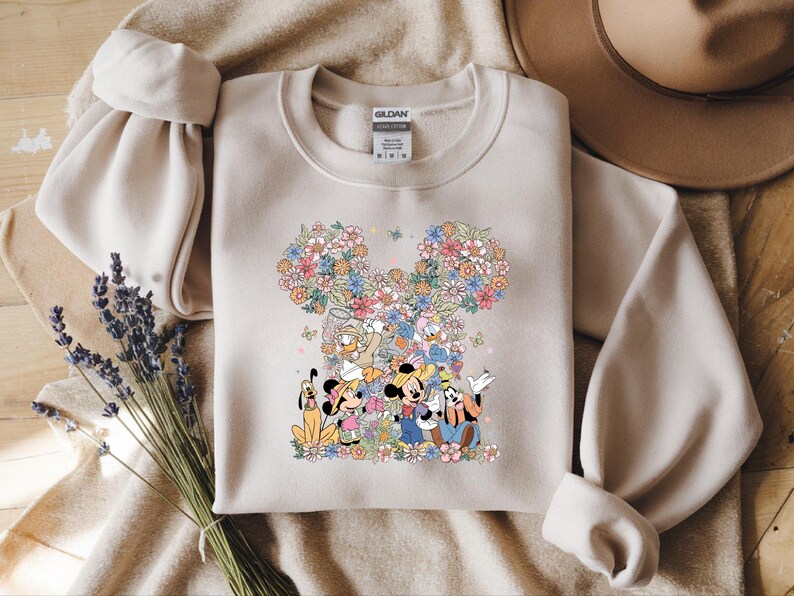 May include: Beige sweatshirt featuring a floral Mickey Mouse design with Mickey, Minnie, Donald, Daisy, Goofy, and Pluto. The design is surrounded by colorful flowers. The sweatshirt has long sleeves and a crew neck.