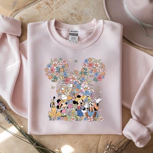May include: A light pink sweatshirt featuring a floral Mickey Mouse design with Mickey, Minnie, Donald, Daisy, and Goofy. The design is surrounded by colorful flowers. The sweatshirt has long sleeves and a crew neck.