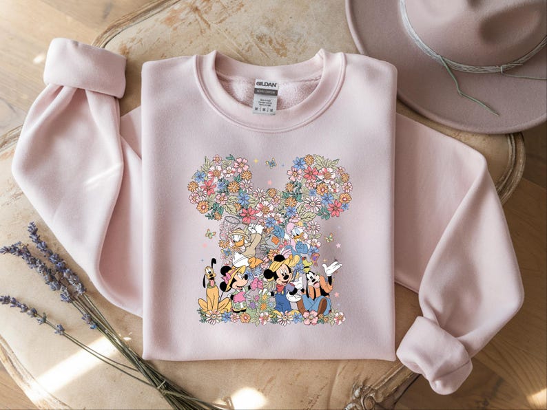 May include: A light pink sweatshirt featuring a floral Mickey Mouse design with Mickey, Minnie, Donald, Daisy, and Goofy. The design is surrounded by colorful flowers. The sweatshirt has long sleeves and a crew neck.