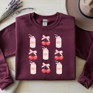 May include: Burgundy crewneck sweatshirt featuring a repeating pattern of Dr. Pepper cans and cherries with pink bows. The Dr. Pepper cans have a straw and whipped cream. A hat and lavender sprig are in the background.