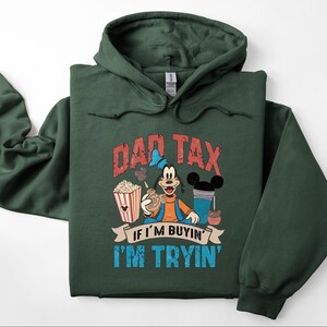 Pode incluir: Uma camisola com capuz verde-floresta escuro com um gr&aacute;fico da personagem Pateta a segurar um snack. O texto DAD TAX IF I'M BUYIN' I'M TRYIN' est&aacute; escrito em letras vermelhas e azuis desgastadas, acompanhado por ilustra&ccedil;&otilde;es de pipocas e uma bebida.