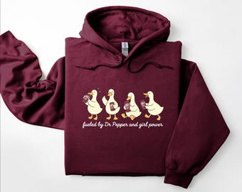 May include: A maroon hooded sweatshirt featuring a graphic of four white ducks holding soda cans. Below the ducks, the text reads, "fueled by Dr Pepper and girl power" in a white script font. The hoodie has long sleeves and a drawstring hood.