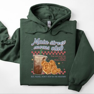 Op de afbeelding: Een donkergroene hoodie met de tekst "Main street moms club". Het ontwerp bevat een koffiekopje, Mickey Mouse wafels en de tekst "Stop and smell the Coffee & Mickey Waffles". De tekst "BUT, PLEASE, DON'T STOP IN THE WALKWAY" is ook zichtbaar.