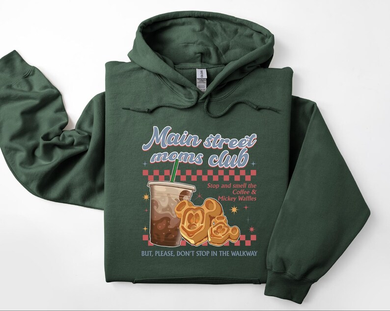 Op de afbeelding: Een donkergroene hoodie met de tekst "Main street moms club". Het ontwerp bevat een koffiekopje, Mickey Mouse wafels en de tekst "Stop and smell the Coffee & Mickey Waffles". De tekst "BUT, PLEASE, DON'T STOP IN THE WALKWAY" is ook zichtbaar.
