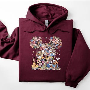 May include: A maroon hoodie featuring a floral Mickey Mouse design. The design includes Mickey, Minnie, Donald, Daisy, Goofy, and Pluto, surrounded by colorful flowers and butterflies. The hoodie has a drawstring hood and long sleeves.