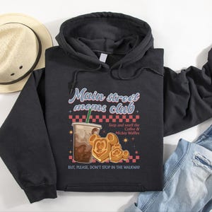Op de afbeelding: Zwarte hoodie met de tekst "Main street moms club" en een illustratie van koffie en Mickey Mouse wafels. De hoodie heeft ook de tekst "Stop and smell the Coffee & Mickey Waffles" en "BUT, PLEASE, DON'T STOP IN THE WALKWAY".