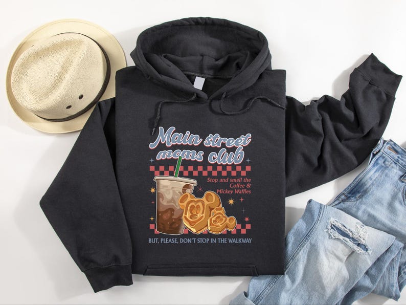 Op de afbeelding: Zwarte hoodie met de tekst "Main street moms club" en een illustratie van koffie en Mickey Mouse wafels. De hoodie heeft ook de tekst "Stop and smell the Coffee & Mickey Waffles" en "BUT, PLEASE, DON'T STOP IN THE WALKWAY".