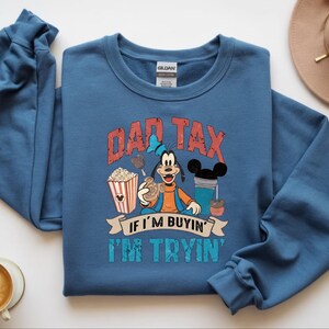 Pode incluir: Uma camisola azul de gola redonda com um gr&aacute;fico do Pateta rodeado por pipocas, uma bebida e snacks. O texto DAD TAX IF I'M BUYIN' I'M TRYIN' est&aacute; impresso em letras vermelhas e turquesa com aspeto desgastado. A pe&ccedil;a tem mangas compridas.