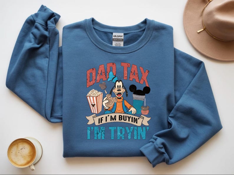 Pode incluir: Uma camisola azul de gola redonda com um gr&aacute;fico do Pateta rodeado por pipocas, uma bebida e snacks. O texto DAD TAX IF I'M BUYIN' I'M TRYIN' est&aacute; impresso em letras vermelhas e turquesa com aspeto desgastado. A pe&ccedil;a tem mangas compridas.