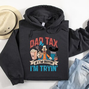 Pode incluir: Uma camisola preta com capuz apresentando um gr&aacute;fico da personagem Pateta rodeado por pipocas, uma bebida e snacks. O texto DAD TAX IF I'M BUYIN' I'M TRYIN' est&aacute; escrito em letras vermelhas e azuis. A camisola tem mangas compridas e um bolso.