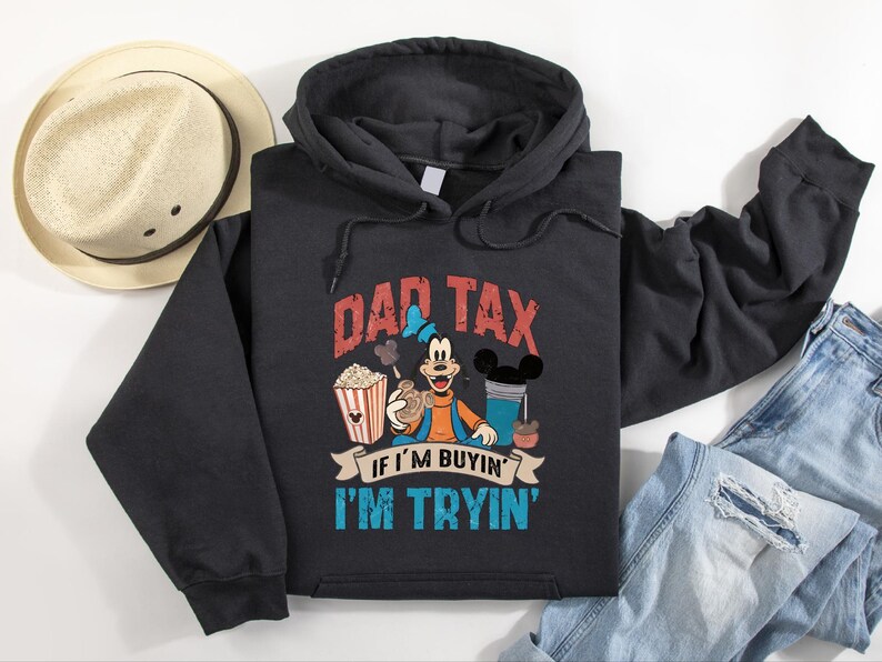 Pode incluir: Uma camisola preta com capuz apresentando um gr&aacute;fico da personagem Pateta rodeado por pipocas, uma bebida e snacks. O texto DAD TAX IF I'M BUYIN' I'M TRYIN' est&aacute; escrito em letras vermelhas e azuis. A camisola tem mangas compridas e um bolso.