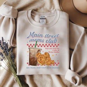 Op de afbeelding: Een beige sweatshirt met de tekst "Main street moms club" en een illustratie van een koffiedrank en Mickey Mouse wafels. De tekst "Stop and smell the Coffee & Mickey Waffles" is ook opgenomen.