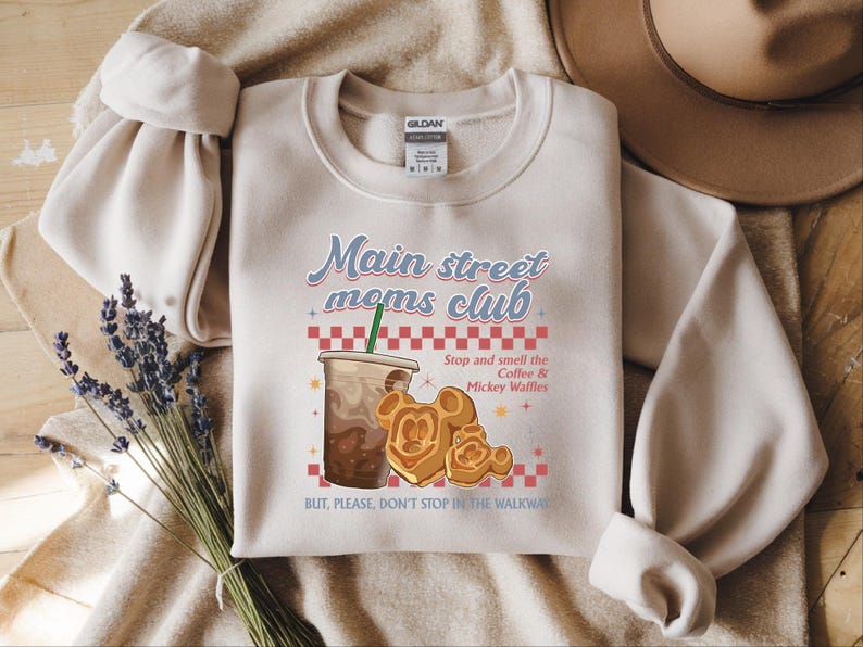 Op de afbeelding: Een beige sweatshirt met de tekst "Main street moms club" en een illustratie van een koffiedrank en Mickey Mouse wafels. De tekst "Stop and smell the Coffee & Mickey Waffles" is ook opgenomen.