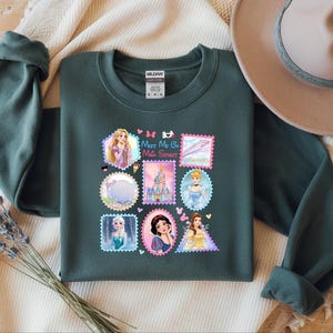 May include: Dark green sweatshirt featuring a colorful graphic of various Disney princesses designed as postage stamps. The text "Meet Me On Main Street" is also included. The sweatshirt has long sleeves and a crew neck.